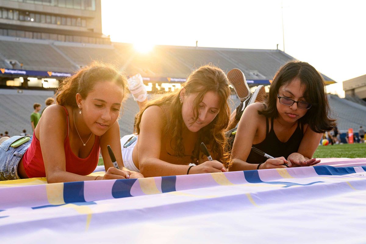 Three students lie on their stomachs and sign a banner on Mountaineer Field.